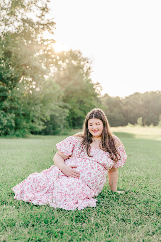 Pregnant woman sitting in the grass with the sun setting behind her as she cradles her baby bump and looks down at it, smiling.