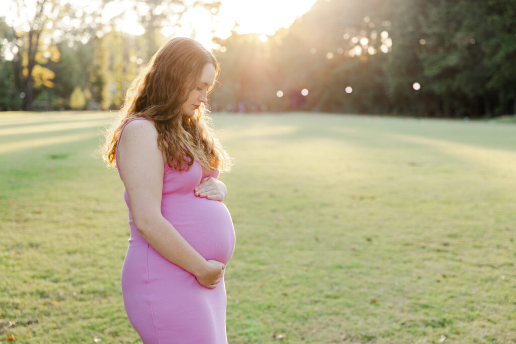 Pregnant girl cradles her baby bump and looks down with a soft smile with the sun setting behind her giving a soft glow