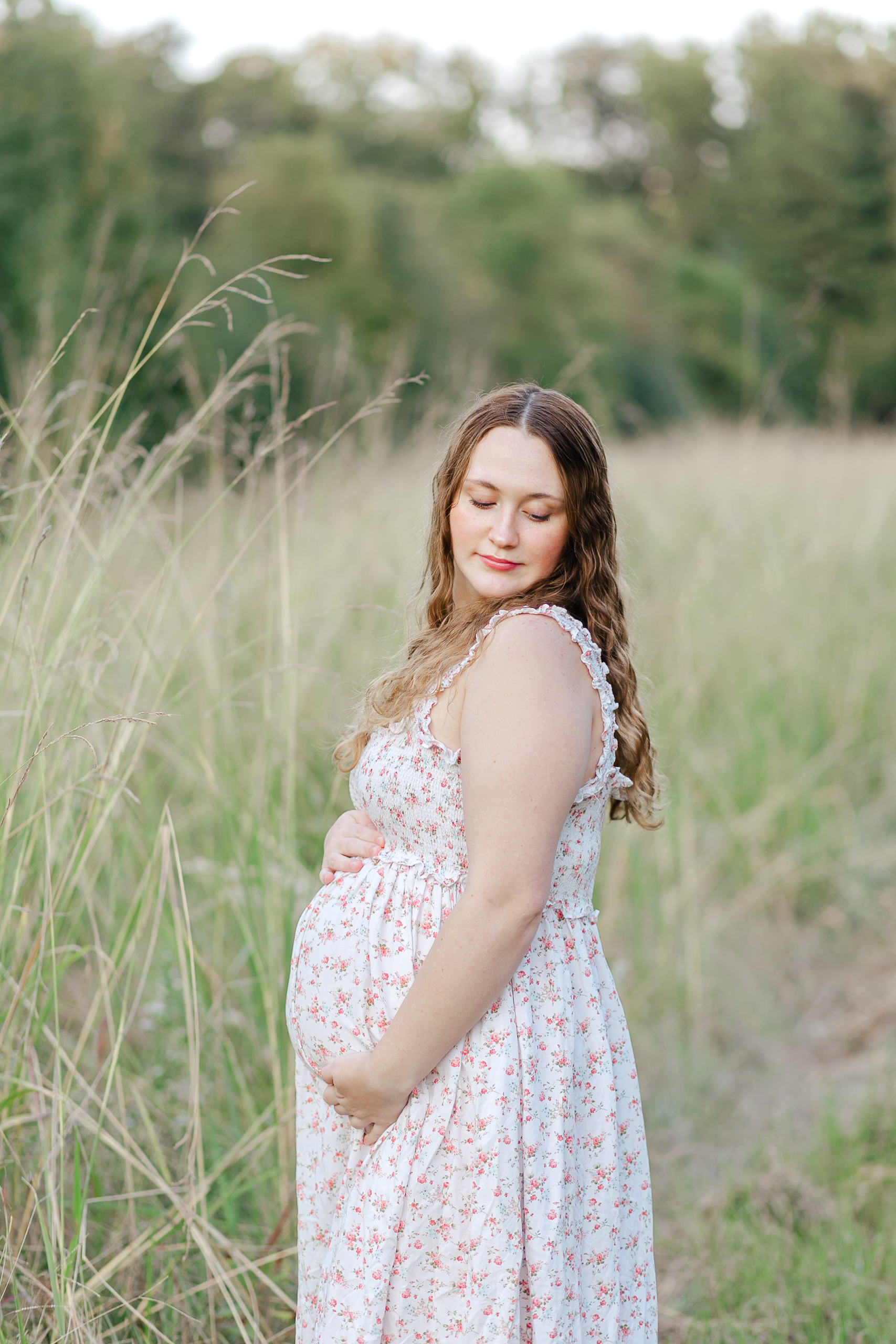 Pregnant woman in a floral dress standing in a serene grassy field, softly cradling her baby bump and gazing downward with a peaceful expression. Captured during golden hour to highlight the natural beauty of motherhood.