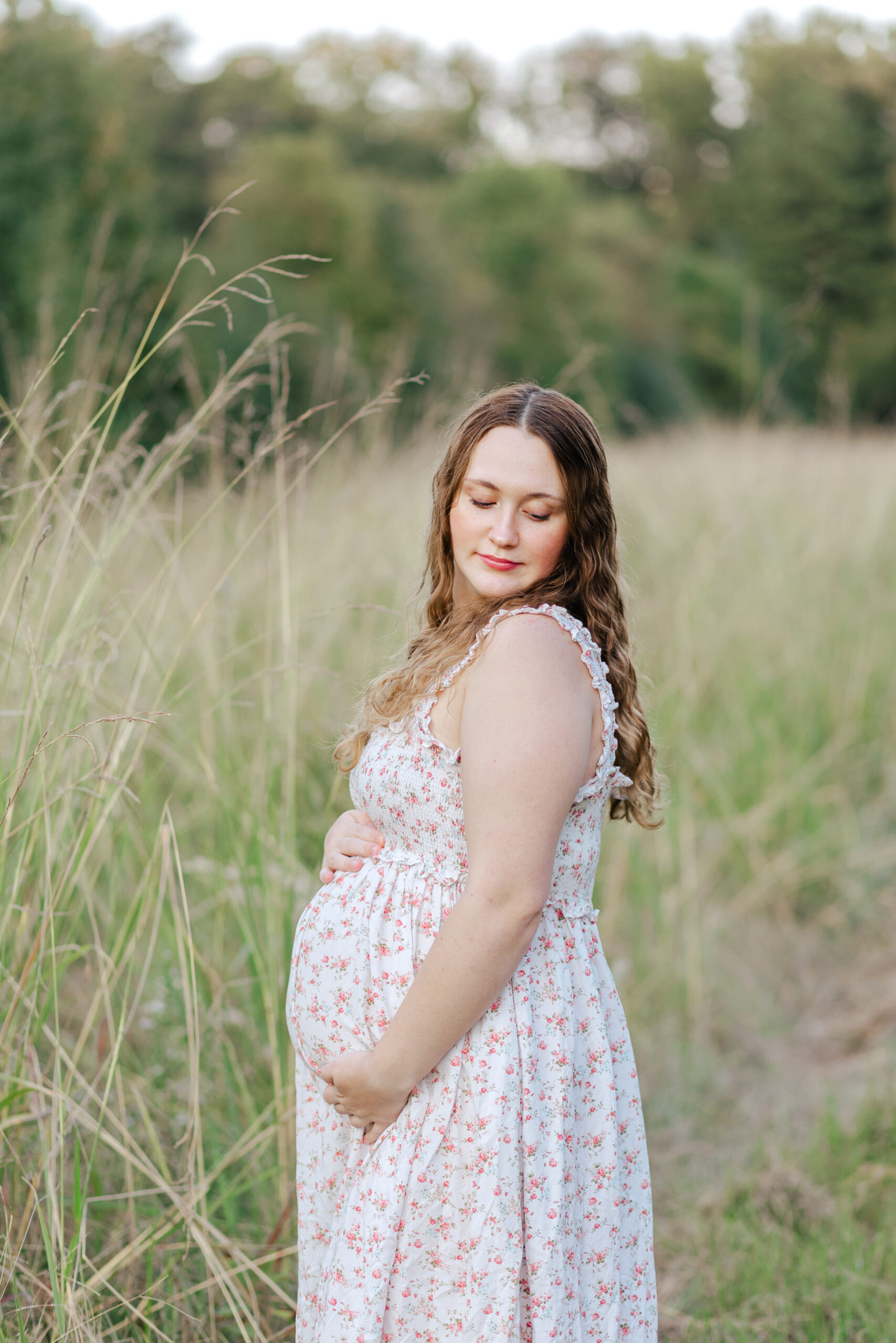 Pregnant woman standing in tall grass and cradling her baby bump and looking down with a soft smile