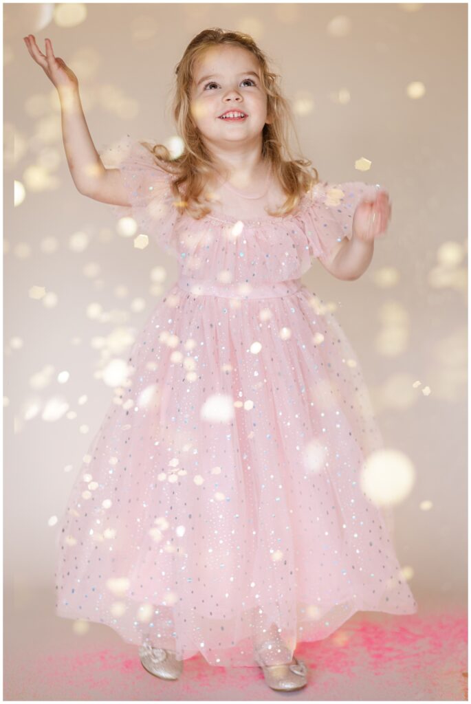 Little girl in a pink dress looking up and smiling as glitter falls down for a glitter photoshoot