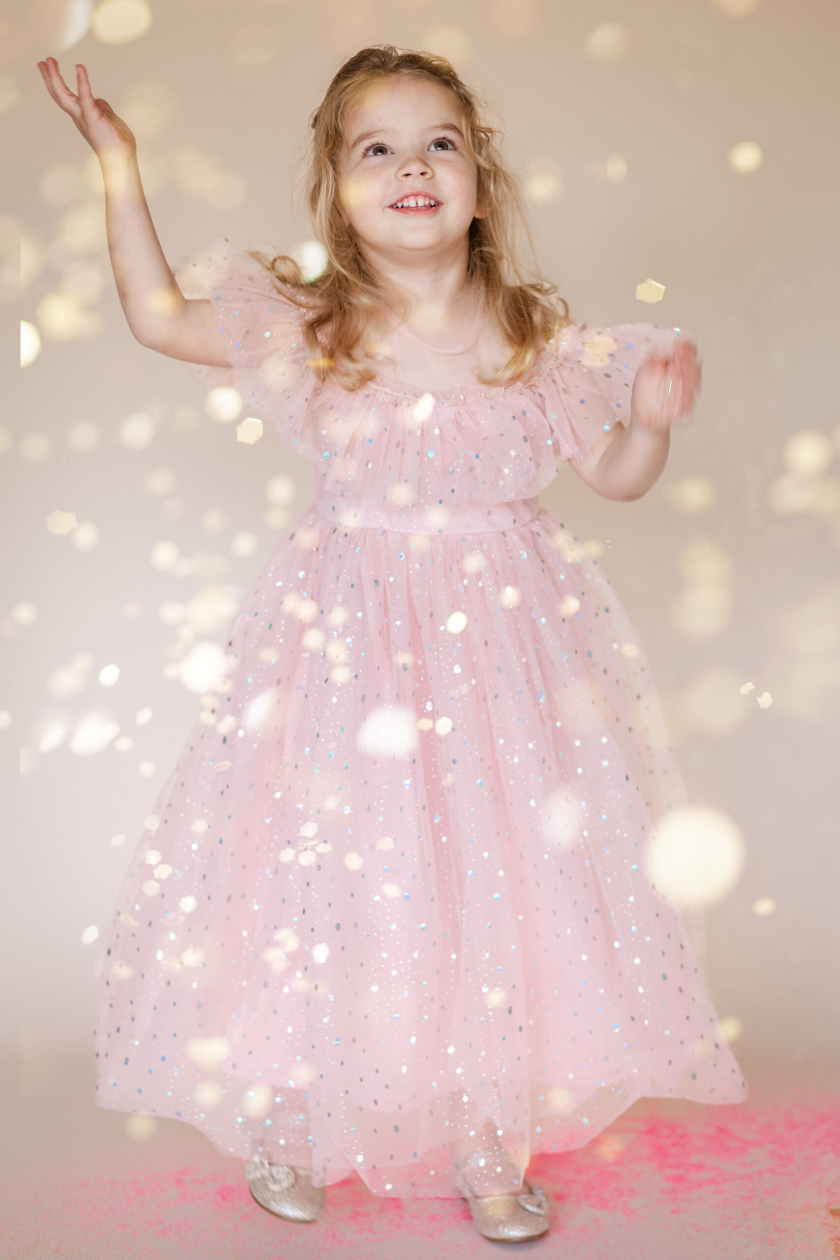 Little girl in a pink dress looking up and smiling as glitter falls down in a glitter photoshoot