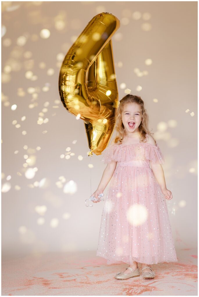 Little girl in a pink dress looking at the camera and laughing while she holds a large number 4 balloon as glitter falls down for a glitter birthday photoshoot 