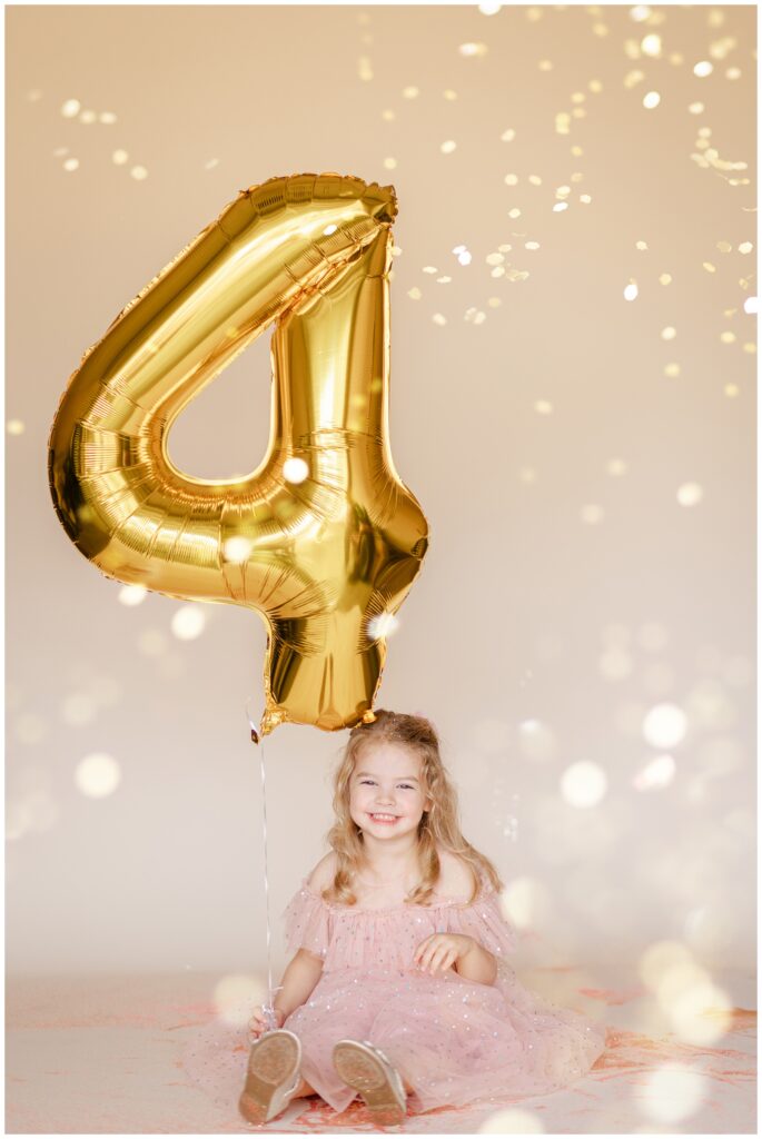 Little girl in a pink dress looking at the camera and laughing while she holds a large number 4 balloon as glitter falls down 