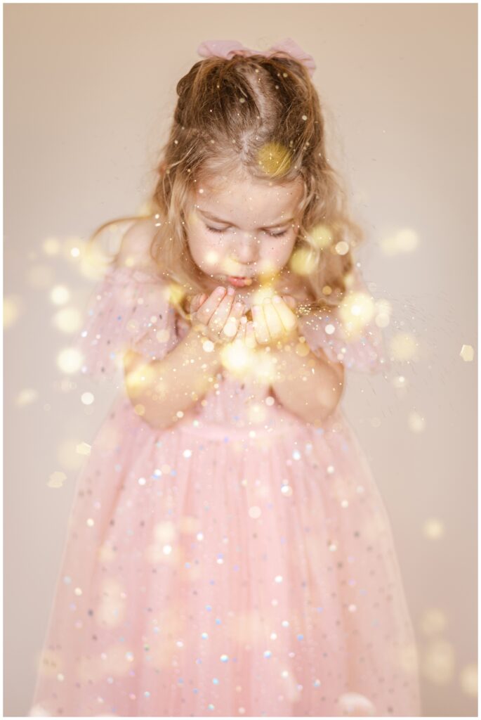 Little girl in a pink dress blowing glitter from her cupped hands towards the camera for a glitter photoshoot