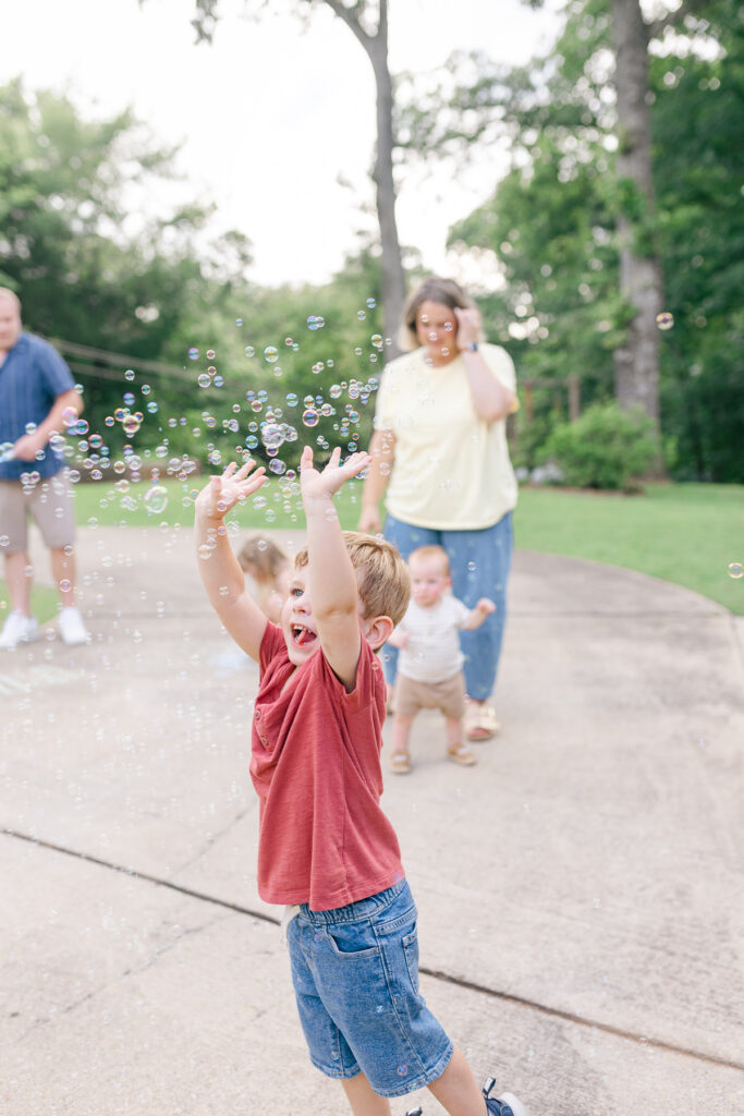 A little boy in a red shirt reaching both hands up to catch a cloud of bubbles.