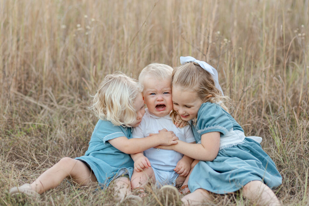 A older sister and brother hugging and comforting their crying little brother in a field, a candid moment from a family photo session in Tuscaloosa
